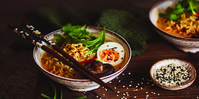 A bowl of ramen noodles with half a hard-boiled egg and cilantro in a small white bowl with brown chopsticks resting on top.